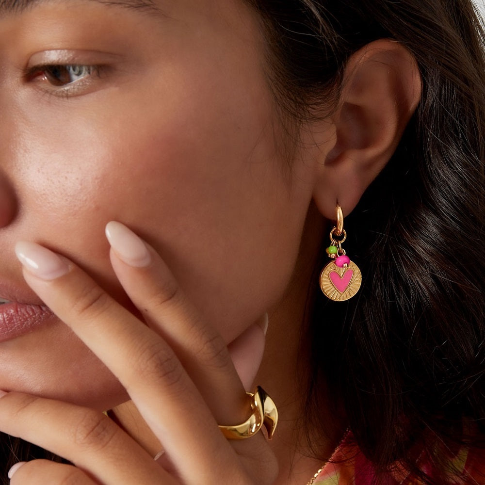 Jonge vrouw met gouden oorbellen. De vrouw draagt de stainless steel oorbellen goud met roze details. De oorbellen hebben een grote bedel met een roze hartje en meerdere gekleurde kraaltjes. De oorbellen zijn waterproof, stainless steel en nikkelvrij. Deze nikkelvrije oorbellen zijn te bestellen op www.jenelry.com